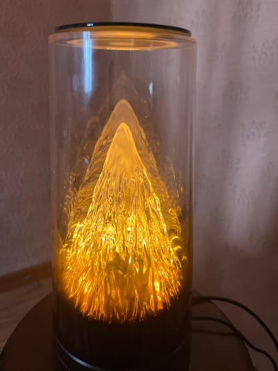A lamp with a mountain-shaped translucent diffuser is positioned on a circular base. The diffuser glows with a warm, golden light, creating illuminated mountain peaks. The lamp is encased in a clear cylindrical glass. The background consists of textured wallpaper. A black electrical cord is visible to the right of the lamp. The image appears to have been taken indoors.
