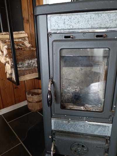 The image shows a close-up of a wood-burning stove in a room with wooden walls and black tiled flooring. To the left of the stove, a stack of firewood is neatly arranged in a black metal holder. Beside the firewood holder, a rustic wooden bucket sits on the floor. The stove itself is dark gray with a glass door that reveals the inside of the firebox, which appears to contain ash and residue from previous fires. The handle of the stove door is black and metallic. Above the glass, there are metal accents with a slightly textured, speckled appearance. Two small, round knobs are visible just above the glass door. The scene suggests a cozy, domestic setting, likely in a home or cabin, possibly in a colder climate given the presence of firewood. The lighting is neutral, with no strong indicators of the time of day. No specific weather conditions are discernible. The brand name "NORDICA" is visible in white text on the lower part of the stove.
