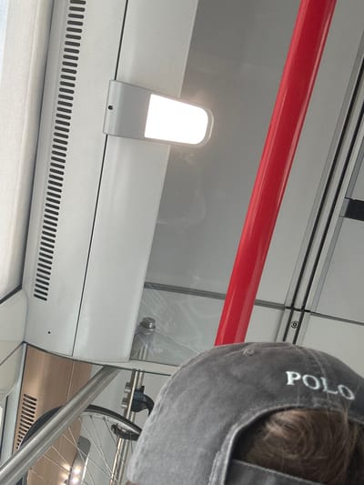 The image depicts an interior view, likely within a public transportation vehicle. A person wearing a dark grey baseball cap with the white embroidered text "POLO" on the back is partially visible in the foreground, their head and shoulder occupying the lower right portion of the frame. The cap is angled downwards, obscuring the wearer's face.
Dominating the mid-ground is a bright, rectangular light fixture mounted on a white panel. To the right of the light fixture and extending vertically through the frame is a vibrant red pole. Beyond these elements, the environment appears to be a modern interior with grey walls and horizontal paneling, suggesting a contemporary design. Reflections are visible on a glass surface, indicating transparency or mirrored elements within the space.
In the lower left quadrant, a bicycle wheel with silver spokes and a black tire is visible, suggesting the presence of a bicycle or bicycle rack. The overall scene is clean and orderly, characteristic of public transit infrastructure.
There is no visual information within the image that would allow for the determination of its location within the city of Salses-le-Château, France. The description is based solely on the visual content provided.