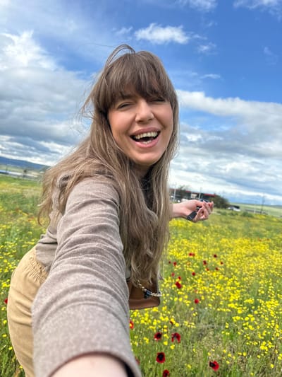 A woman with long, light brown hair and bangs smiles widely, revealing her teeth, in a selfie taken in a field of yellow and red flowers under a partly cloudy blue sky. She is wearing a long-sleeved beige shirt and khaki pants. Her right arm is outstretched, holding a small black object. A brown purse with a gold chain strap is visible on her right side. In the background, there are power lines, some trees, and a road with a vehicle. The location is likely in the Marneuli region of Georgia, based on the distinctive landscape of flowering fields and rolling hills.