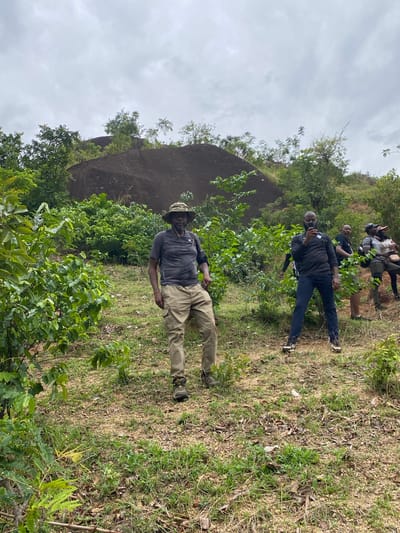 A photograph shows a group of people outdoors, likely in a rural or natural setting. The foreground features dense green vegetation and grassy terrain. A man in a hat, gray shirt, and khaki pants is in the mid-ground, closer to the camera. Behind him, two other men stand closer to a rock formation. A large rock formation is behind the vegetation. The sky is cloudy. The photograph may have been taken in Ganawuri, Nigeria.