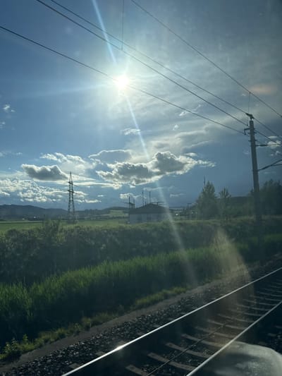 The image shows a view from a moving train in Loosdorf, Austria. The foreground features railroad tracks, with ballast and ties visible. Adjacent to the tracks is a grassy embankment. In the midground, a field of green grass stretches towards the horizon, with a power line tower in the distance. Several structures, including what appear to be industrial buildings, are also present. A line of trees and rolling hills form the background. The sky is a mix of blue with scattered white and gray clouds. Bright sunlight streams down, creating lens flare effects. The overall impression is of a rural landscape viewed from a train.