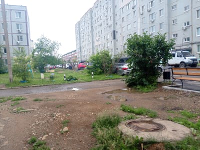 The image depicts a residential courtyard in Arseniev, Russia. A multi-story apartment building with a light gray facade and numerous windows dominates the background. In front of the building, several parked cars, including a white minivan with roof racks and a red sedan, are visible on a paved surface. Green trees and bushes are scattered throughout the courtyard, with a playground structure partially visible on the left. The foreground shows a dirt and gravel area with patches of grass and a manhole cover. Puddles of water are present on the paved area and the dirt surface, suggesting recent rain. A wooden bench with a metal frame is positioned to the right. The sky is overcast.
