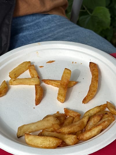A close-up shot shows a white paper plate on a red surface, with several french fries arranged to form the number "4" and a sideways "J" or "1" character. The fries are golden brown and appear to be slightly greasy. In the background, out of focus, a person is visible wearing blue denim jeans and a brown garment. The scene is outdoors, indicated by the green foliage in the upper right corner. The context suggests a casual outdoor meal. The specific location in Sainte-Marie-au-Bosc, France, is not discernible from the visual information provided in the image.