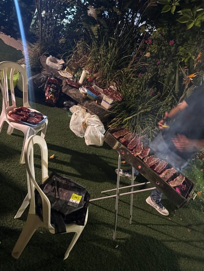 The image depicts an outdoor night-time barbecue scene in a residential backyard or patio in Kfar Yona, Israel. The ground is covered with artificial green turf.
A long, rectangular charcoal grill, oriented diagonally from the lower right to the upper middle, is actively in use. Approximately ten to twelve cuts of raw, seasoned red meat, consistent with steaks or ribs, are arranged across the grill's grates. Smoke is visibly rising from the cooking meat.
A person is partially visible to the right of the grill, wearing dark clothing, blue jeans, and white sneakers with black accents. Their hands are positioned over the grill, suggesting they are tending to the cooking process.
To the left and slightly behind the grill, a low, dark-colored surface or wall serves as a food preparation area, backed by dense green foliage. This area holds various containers and trays of raw meat, along with other dishes, utensils, and a large bag of charcoal or briquettes.
Two white plastic chairs are present. The chair in the mid-left holds a blue tray filled with multiple pieces of raw, red meat. The chair in the lower-middle-left has a black bag with a yellow label resting on its seat.
Several full, white plastic bags are situated on the turf between the chairs and the food preparation area. The background features dense, dark green foliage with some visible pink flowers and a "Bird of Paradise" bloom, illuminated by an artificial light source, creating shadows and highlighting the active grilling environment.