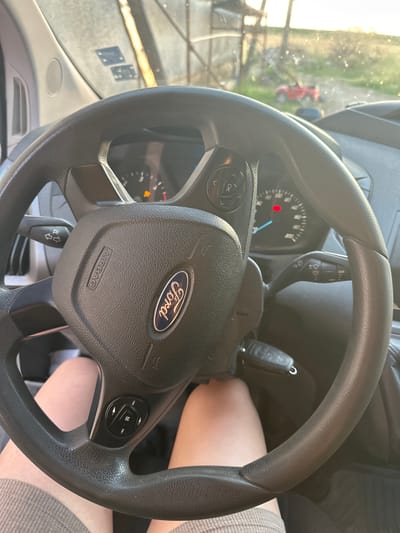 The media file is a close-up, interior shot of a Ford vehicle's steering wheel and dashboard. The steering wheel is black and features the Ford logo prominently in the center. Below the logo, the word "AIRBAG" is visible. On the left side of the steering wheel, there are buttons with directional arrows and an "OK" button. On the right side, there is a circular control with buttons labeled "M", and forward/backward arrows, likely for media controls. A car key is inserted into the ignition on the right side of the steering column.
The dashboard includes a speedometer with a red warning light illuminated and an orange indicator light. The windshield offers a blurred view of an outdoor scene, with greenery and a red vehicle visible in the distance. Sunlight streams in, suggesting it is daytime, and the weather appears clear. The overall scene depicts the interior of a vehicle, possibly a van or truck, ready for operation. No people are clearly visible, though a person's legs are partially visible beneath the steering wheel. The setting appears to be a parking area or roadside, given the view through the windshield. No specific text beyond the visible labels on the steering wheel and dashboard is discernible. The context provided indicates the location is Mlekarevo, Bulgaria.