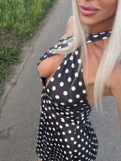 The image depicts a close-up, partial view of a person outdoors, rotated 90 degrees clockwise. The subject has long, platinum blonde hair and is wearing a black garment adorned with white polka dots, possibly a dress or a top with thin straps, which exposes cleavage. A golden hoop earring is visible on their right side, near the jawline. Their face is partially shown from the nose down, highlighting full, light pink lips. A small, cursive tattoo, which appears to read "Jack" or a similar name, is visible on their upper chest/collarbone area.
The setting is outdoors during daylight hours, characterized by clear ambient lighting. The person is positioned on a paved surface, likely asphalt or concrete, with visible aggregate. Alongside the paved area, a strip of green grass and various weeds grows, suggesting a roadside, pathway, or park environment. The photograph is taken from a slightly elevated perspective, looking down at the subject. The person appears to be casually posing for the camera. No other individuals or significant objects are present in the frame. The location context provided is Конь, Russia.