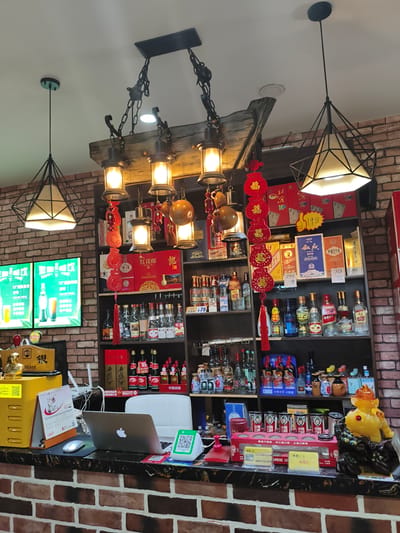 The image depicts the interior of a shop, possibly a liquor store or convenience store, located in Jixi, China. The scene is set against a backdrop of brick walls, with shelves stocked with various bottles of alcohol and boxed goods. Decorative elements like red Chinese lanterns and traditional decorative gourds hang from a rustic wooden beam holding multiple light fixtures.

In the foreground, a counter displays a laptop, a computer mouse, a scanner, a calendar, and a yellow cash register. A QR code payment terminal is also visible on the counter. A statue of a golden mythical creature, possibly a lion or dog, is placed on the right side of the counter. The lighting appears to be a mix of overhead and directional lamps, creating a warm ambiance. There are no people clearly visible in the image, though the setup suggests a retail environment where a cashier would operate. The overall impression is one of a well-stocked and decorated shop in a Chinese urban setting.
