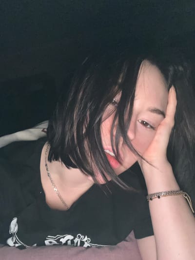 A woman with dark hair partly obscuring her face is shown close up. She is wearing a black t-shirt with a white design. Her hand is resting on her forehead. She has a silver chain necklace and bracelets on her wrist. The background is dark.