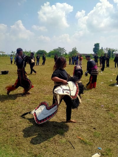 This image captures an outdoor cultural performance or practice session in Celep, Indonesia, under a bright, partly cloudy sky. The scene is set on a large, open field of dry, sparse green and brown grass, with lines of trees and vegetation visible along the distant background.
In the mid-ground, a female participant with long, dark brown hair, barefoot, wearing a black t-shirt and black leggings, holds a flat, white, horse-shaped prop (kuda lumping) decorated with red and black patterns and a shaggy black mane. She looks to her right. Another similar, larger prop lies partially visible on the ground at her feet, casting a distinct shadow. To her left, a male participant, also barefoot, wears a black long-sleeved shirt and an elaborate patterned sarong or trousers in red, black, and white. He moves towards the right side of the frame, holding a dark, shaggy costume element or prop. A red head covering is visible on him. Further to the right in the mid-ground, another barefoot participant in a black top and a patterned sarong (white, black, red) is bending forward, with a dark, shaggy prop near them.
Approximately 15-20 additional individuals are scattered across the mid-ground and background. Some are dressed similarly to the primary performers in dark attire with traditional elements, while others in the far background appear to be observers wearing casual clothing. A black bucket is visible on the ground in the far left of the frame. Small pieces of litter are scattered on the grass in the foreground. The strong sunlight indicates a daytime setting.
