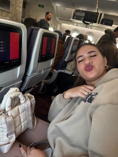 An interior view of a commercial aircraft cabin, likely situated at Mohammed V International Airport (CMN) in Nouaceur, Morocco. The primary subject is a woman in the immediate foreground, positioned facing the camera. She has her eyes closed and lips pursed in a kissing gesture, accentuated by dark pink lipstick. She wears a beige hooded sweatshirt featuring a dark graphic on the chest, small gold hoop earrings, and a large, multi-faceted ring on her right ring finger. Her left hand, adorned with a plain band ring, rests near a quilted, light-colored shoulder bag placed on an adjacent seat. In the background, multiple rows of passenger seats are visible, each equipped with an individual in-flight entertainment screen displaying language options including English, Arabic, French, and Portuguese. Overhead bins are closed, with some luggage discernible inside them. A male passenger with a beard is standing in the aisle towards the rear of the cabin. Other passengers are seated further back. The cabin features blue and purple seat accents, a patterned reddish-brown carpet, and is illuminated by interior lighting.