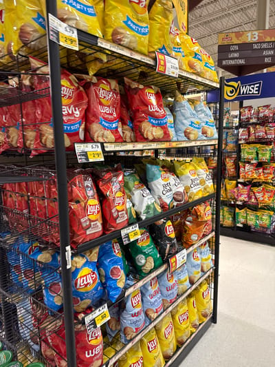 The image displays a potato chip aisle inside a ShopRite supermarket in Lawrence Township, United States. A black, multi-tiered metal wire shelving unit dominates the scene, densely stocked with various brands and flavors of potato chips. Large "Party Size" bags of Lay's Wavy potato chips, predominantly in red packaging, are prominently displayed on the upper and middle shelves, with price tags indicating $5.79 or a sale price of $2.99. Other visible Lay's varieties include Hot Sauce, Sour Cream & Onion, Salt & Vinegar, and Lightly Salted. Kettle Cooked chips, in flavors like Jalapeño and Original, are also present, some priced at $4.49 with a $0.50 discount. To the right, another retail shelving unit is visible, holding additional snack products, with a blue sign bearing the "WISE" brand logo. An overhead brown and white directional sign with the ShopRite logo and the number "13" points to sections labeled "TACO SHELLS / MASAS," "LATINO," "ASIAN," "INTERNATIONAL," and "RICE." The supermarket floor is light-colored. No individuals are present in the image.