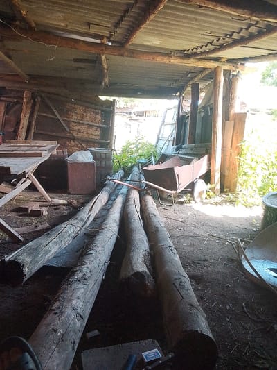 The image shows the interior of a rustic, open-sided structure, likely a shed or workshop, in Izgutty Ajtykov, Kazakhstan. The roof is made of corrugated metal sheets supported by wooden beams, some of which appear weathered and rusted. The walls are partially constructed from wooden planks and sheets of corrugated material. The floor is dirt and debris.
In the foreground, several large, rough-hewn logs are laid out horizontally. To the left, a wooden workbench or table with a hinged top stands on legs. Further back, there is a rusty metal container, possibly a bin, with a white plastic bag inside. Beside it, a wooden barrel stands upright. In the mid-ground, a rusty wheelbarrow with a damaged body is positioned. Beyond the wheelbarrow, there is dense green foliage visible, suggesting the structure is located outdoors or adjacent to a vegetated area. The overall impression is one of a storage or work area for materials, with a utilitarian and somewhat neglected appearance. No people are visible in the image.