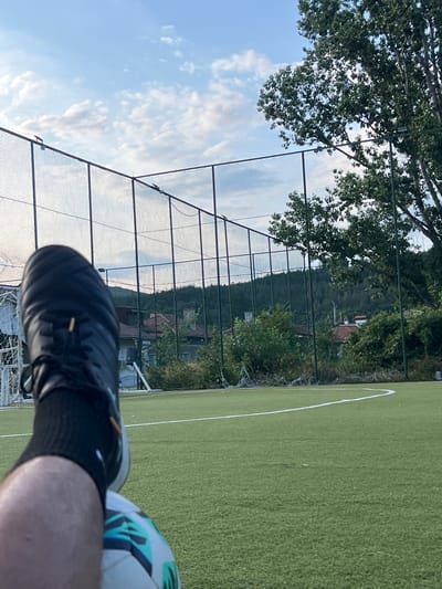 A person's leg, clad in a black sock and a black soccer cleat, rests on a soccer ball on a green artificial turf field. The field is bordered by a chain-link fence, beyond which are trees, houses with red-tiled roofs, and a forested hillside under a cloudy blue sky. The scene appears to be a soccer field located in Belovo, Bulgaria.