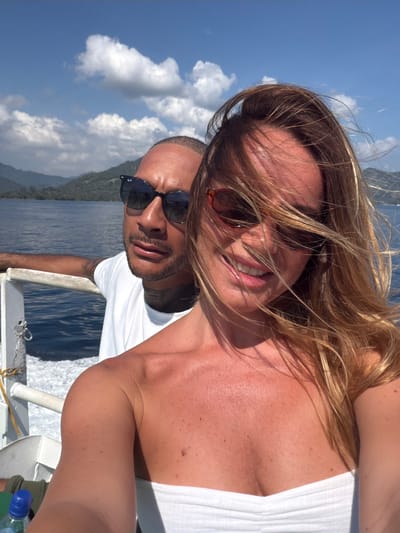 A man and a woman are taking a selfie on a boat in the waters off Pemenang Timur, Indonesia. The man, wearing a white t-shirt and black sunglasses, leans on the boat's railing. The woman, with her hair blowing in the wind and wearing a white strapless top, smiles at the camera. The background shows blue water, green hills, and a cloudy sky.
