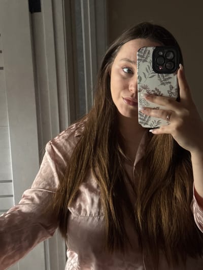The image depicts a mirror selfie of an adult female, visible from the chest up, in an indoor setting in Tlustenkhabl, Russia. The subject has long, dark brown hair and is wearing a light pink, subtly patterned, long-sleeved shirt, possibly satin or silk. She holds a dark-colored smartphone with a light-colored, floral-patterned case in her right hand, obscuring the lower-left portion of her face. A plain metallic band ring is present on the ring finger of her right hand, and her fingernails are manicured with a light polish. Her right eye is visible, appearing light-colored. The background features vertical panels or blinds, predominantly light gray. Strong directional lighting illuminates the right side of the subject's face and creates shadows on the left.