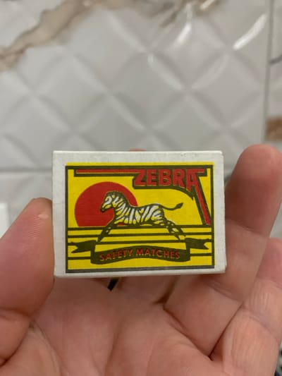 An adult human hand holds a rectangular white matchbox, presenting its front label directly to the viewer. The label features a prominent yellow background with a thick black and red border. Centered on the label is a stylized black and white zebra depicted in a running or leaping posture, superimposed over a large red circle. Above the zebra, the word "ZEBRA" is printed in red capital letters with a black outline. Below the zebra, a black banner contains the text "SAFETY MATCHES" in green capital letters. The hand, visible with skin texture and finger creases, grips the sides and bottom of the matchbox. The background consists of light-colored, possibly off-white, wall tiles with a distinct raised, diamond-shaped pattern and subtle brownish veining, suggestive of marble or natural stone. The scene is located in Balgarsko Slivovo, Bulgaria.