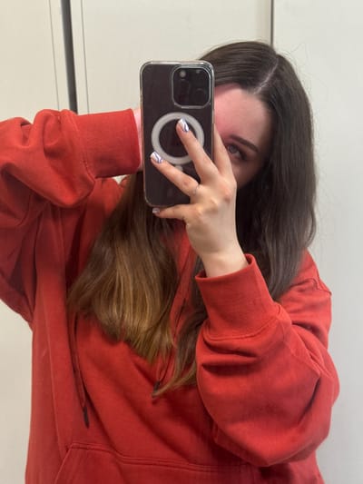 In a room with plain, off-white walls, a person is taking a selfie in a mirror. The person is wearing a bright red hoodie with the hood up and is holding a smartphone in their right hand to take the photo. The phone has a clear case with a silver ring accessory. The person's left hand is partially visible, resting near their face. Their dark hair is visible on the sides of their head, and a portion of their face, including an eye and eyebrow, is visible. The lighting suggests an indoor setting. The image does not contain enough information to determine the specific location within Гамово, Russia.
