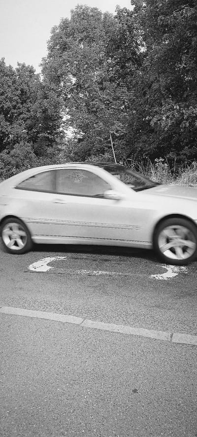 A black and white, medium shot captures a silver coupe driving on a paved road, with a line of trees bordering the right side of the frame. The car is in motion, evidenced by the blur of its wheels and the slight streaking of its body. A white, curved arrow painted on the road indicates a turn or lane change. The trees in the background appear lush and dense, with some grass and weeds growing in front of them. The sky is overcast and washed out. This scene is located on Hauptstraße in Waiblingen, Germany.