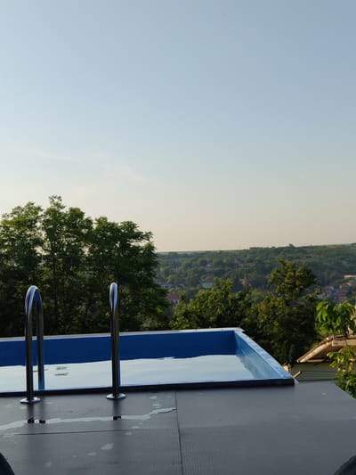 The scene is set outdoors in Pisanets, Bulgaria. A rectangular swimming pool with a blue interior, partially filled with clear water, occupies the central foreground. Two curved, metallic (likely stainless steel) ladder handrails are anchored to the dark gray, textured surface surrounding the pool. Small puddles of water are present on this surface near the ladder. Beyond the pool, a dense expanse of various green trees dominates the mid-ground, obscuring parts of a distant village or town. This settlement is characterized by structures with light-colored walls and distinct reddish-orange roofs, nestled among rolling green hills. The background features a clear, expansive sky, transitioning from a light blue overhead to a paler, almost yellowish-white hue at the horizon. No individuals or specific actions are discernible within the frame. Indistinct dark shapes are partially visible at the extreme bottom edges of the image.