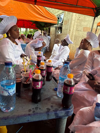 In Ikoga, Nigeria, a gathering is taking place under an orange tent. Several individuals, mostly women, are seated. They are dressed in white attire, with many wearing white, pleated headwear. In the foreground, a blue plastic table is set with several plastic cups containing dark liquid and colorful straws. There are also bottles of water and soda on the table. The people appear to be in a relaxed, social setting, possibly at an event or ceremony. The background shows a rough-textured building.