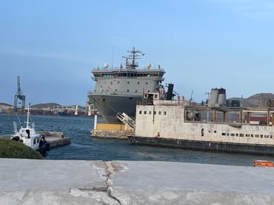 In Cartagena, Spain, a large grey naval vessel is docked alongside a derelict white ship. The naval vessel has several antennas and navigation equipment on its bridge, and small flags are flying from its superstructure. The white ship appears weathered and rusted, with visible signs of decay. In the foreground, a small white tugboat is moored to a stone jetty where two individuals are standing. A rough concrete pier occupies the immediate foreground. In the background, cargo ships and cranes are visible in the harbor, indicating an active port. The sky is clear and light blue.