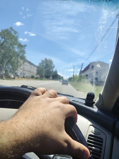 The image shows a person's hand on a steering wheel, driving on a road in Shimanovsk, Russia. The sky is blue with scattered clouds. Trees and buildings line the road. A small, dark figurine is visible on the dashboard. The windshield has several smudges and streaks.
