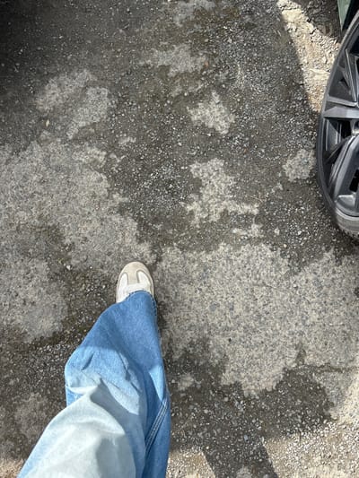 The image shows a person's right leg wearing blue jeans and a white sneaker, standing on a gravel driveway in Dinard, France, at the coordinates 48.631, -2.054. The gravel is a mix of light gray and dark gray, with patches of a lighter material that may be sand or crushed stone. The image is taken from a first-person perspective, looking down at the leg. The background shows the tire of a black car to the right of the image. The person's shadow is visible behind the leg.