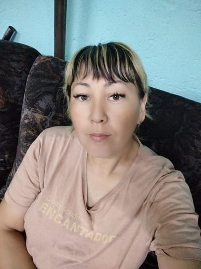 The image shows a woman with blonde hair with dark streaks, bangs, and visible makeup. She is wearing a light pink t-shirt with the Spanish words "Complicadamente ENCANTADO" printed in a lighter shade. The woman is seated on a dark, patterned sofa. Behind her is a light blue wall. The scene appears to be indoors, likely in a residential setting. The location of Зелёное, Russia is not depicted or suggested by any elements in the image.