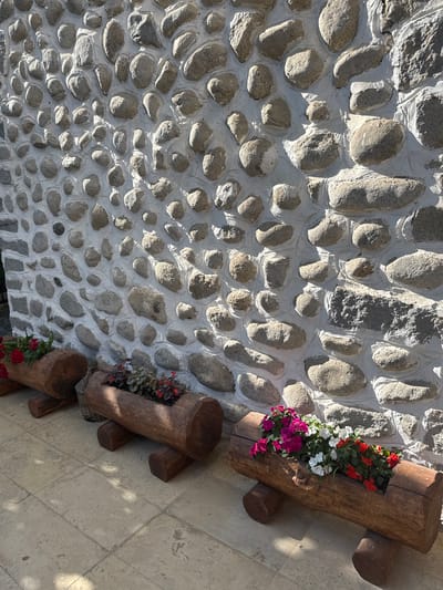 The image depicts an outdoor architectural detail in Melnik, Bulgaria, showcasing a textured stone wall fronted by three wooden log planters on a tiled ground. The wall is constructed from numerous irregularly shaped, light grey stones of varying sizes, deeply embedded in thick white mortar. The stones protrude significantly from the wall surface, casting distinct, sharp shadows under direct sunlight, emphasizing the wall's rugged texture. An arched opening, also composed of stone, is visible on the far left. A section towards the upper right appears to incorporate larger, flatter, rougher-textured grey stones.

Three hollowed-out wooden logs, functioning as planters, are positioned in a linear arrangement along the base of the wall. Each log is supported by two smaller, cylindrical log segments serving as feet. The leftmost planter contains red flowering plants. The middle planter features smaller red flowers and dark, possibly burgundy, foliage plants; a woven wicker basket is partially visible behind it. The rightmost planter displays a vibrant assortment of pink, white, and red flowers interspersed with green foliage.

The ground is paved with large, rectangular, light-colored flagstones or tiles, exhibiting minor textural variations and patches of shadow from an unseen overhead source, contrasting with areas illuminated by direct sunlight. No individuals are present in the scene.