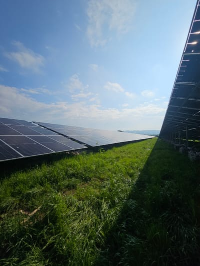 The image depicts a solar farm in Lacock, United Kingdom. Parallel rows of solar panels stretch into the distance under a partly cloudy blue sky. To the right of the panels is a field of lush green grass. The panels appear clean and well-maintained.