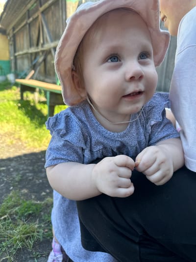 A baby with blue eyes and blonde hair, wearing a pink hat and a blue dress, is held by an adult in a white shirt. The baby is looking up and to the right, with a slight smile. The background shows a wooden fence and a bench. The location is Novoyeniseyka, Russia. The image is taken outdoors during the day.