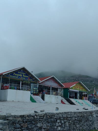 The image depicts a row of dhabas, or roadside eateries, situated at the Marhi location in India, nestled against a misty mountainside. The dhabas are brightly colored, with red-tiled roofs and signs indicating their names and offerings. The central dhaba, "Himalayan Rasoi Punjabi," prominently displays "Height 11200 feet" and advertises "Breakfast, Lunch, Snacks." To its right, "Friends Fouji Dhaba" also advertises similar fare. Further to the right is "Valley View Dhaba," again highlighting "Breakfast, Lunch, Snacks" and noting its height as "11,200 Ft." The dhabas have outdoor seating areas and stairs leading up to them, some of which are painted green and white. In the foreground, a stone wall runs along the lower edge of the image. The environment is overcast and hazy, with the mountains in the background shrouded in mist, creating a sense of elevated altitude. A few people are visible inside or near the dhabas, and a van is parked to the right of the "Valley View Dhaba."
