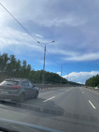 The image shows a view from inside a car, looking out onto a multi-lane highway in Новосёлки, Russia. A gray SUV is visible in the adjacent lane, and other cars are seen further ahead on the highway. The highway is lined with streetlights and bordered by trees. The sky is blue with scattered white clouds. The photo appears to have been taken during the daytime. The license plate number of the gray SUV is partially visible as "K 298 EE 52".