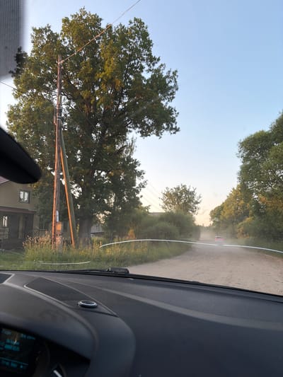 The image captures a view from inside a vehicle, looking out through the windshield. The scene depicts a rural road in the city of Nelyudovo, Russia. The road appears to be unpaved and dusty, with a single car visible in the distance creating a dust trail. On the left side of the road, there is a house with a porch and a large tree next to a utility pole. The sky is clear and light blue, suggesting it is daytime. The foreground shows the car's dashboard.