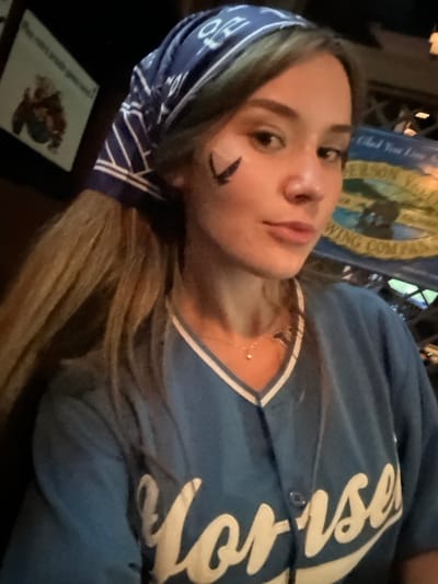 A young female subject is depicted in an indoor, dimly lit environment, likely a bar or pub, in 서대문구, South Korea. She wears a blue bandana with white patterns covering part of her long, light brown hair. A black, stylized bird emblem is painted on her left cheek. She is wearing a light blue baseball-style jersey with white trim around the collar, featuring white embroidered text, partially visible as "Yorise," possibly indicating "Yonsei." A thin necklace with a small, spherical pendant and a secondary charm is visible on her neck. The background includes a dark-framed picture on the left and a larger, lighter sign on the right with partial text "ANDERSON VALLEY BREWING COMPANY." The subject faces the camera directly with a neutral expression.