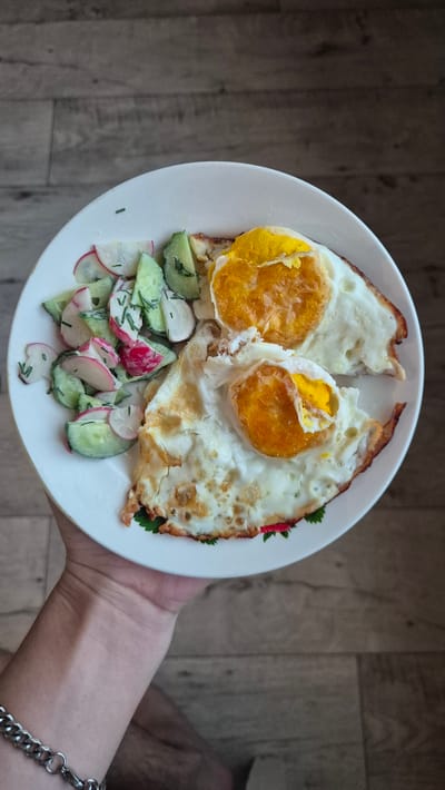 A hand holds a white plate containing two fried eggs and a side salad. The eggs have well-cooked whites and bright yellow yolks. The salad is composed of sliced radishes, cucumber, and dill, lightly dressed. The plate is set on a wooden surface. The image was taken in Bulatovo, Russia.