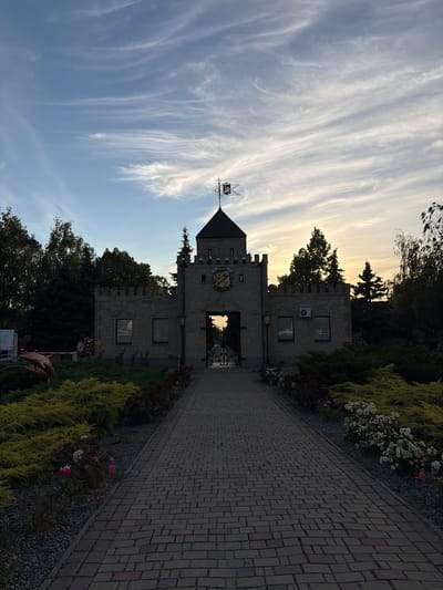 The image captures a twilight view of a castle-like structure with a paved walkway leading towards its entrance in Slobozhanske, Ukraine. The sky displays wispy clouds, with the sun setting or having recently set, casting a warm glow on the horizon. The building, constructed from stone, features crenellated battlements and a central tower topped with a weathervane. In front of the structure, there are manicured bushes, flowers including roses, and a path made of brick pavers. A bronze sculpture of a dolphin is visible to the left of the walkway, situated within a landscaped area. The scene is devoid of people, and the overall atmosphere suggests a tranquil, possibly park-like setting at the end of the day.
