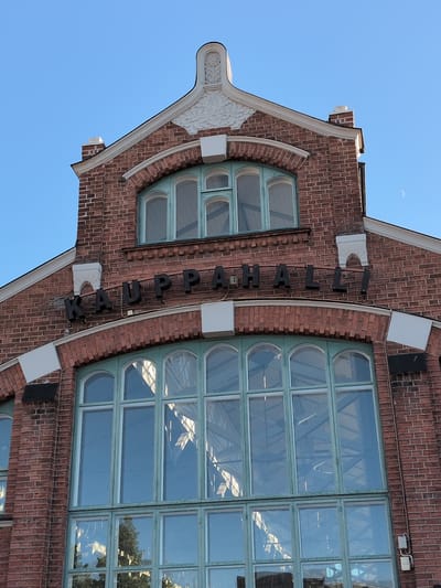 The image shows the facade of the Oulu Market Hall (Oulun Kauppahalli), a historic building located in the city of Oulu, Finland. The facade is constructed of red brick and features large, arched windows with green frames. Above the windows, the word "KAUPPAHALLI" is visible in black, three-dimensional lettering. The sky is clear and blue, with a crescent moon visible in the upper right quadrant of the image. Architectural details, such as decorative white elements above the windows and a chimney, are also present. The overall impression is of a well-maintained, classic building against a bright sky.