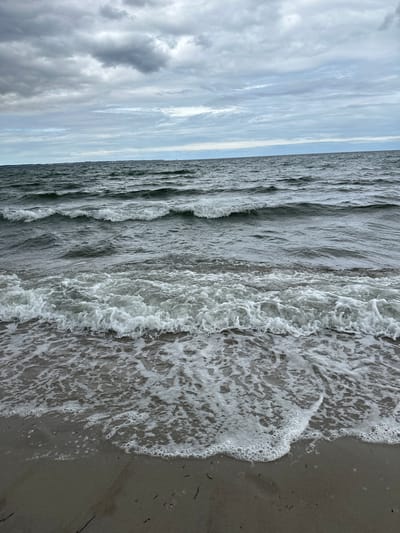 The image shows a view of the Baltic Sea from the beach in Scharbeutz, Germany. The sky is overcast with gray clouds. The sea is choppy with waves breaking on the shore, creating white foam. The beach is made of sand and there are some scattered pieces of seaweed. There are no people or objects visible in the image, except for the natural elements.