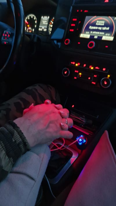 Inside a car at night or in low light, two individuals are holding hands in the center of the frame, illuminated by a warm red glow. The car's interior is visible, featuring a dashboard with illuminated controls and an infotainment system.

The left hand, likely female, has long, light-colored manicured nails and wears a silver-colored ring on the ring finger. It is clasped with a second hand, likely male, which has shorter nails. The person whose hand has the ring wears a light-colored, possibly beige or cream-colored, sleeve, while the other wears a dark, patterned (possibly camouflage) sleeve. The gesture of holding hands suggests affection or companionship.

On the car's infotainment screen, text in Cyrillic is displayed, indicating music playback. The visible text includes "Владимир Адамиров" (Vladimir Adamirov), likely the artist's name, and "Журавли над тайгой" (Cranes over the taiga), which appears to be the song title. Below this, "2:22" indicates elapsed time, and "3:14" is the total song duration. Other labels visible on the screen are "MDI" (Media Device Interface) and "Выбор" (Selection).

The car's climate control panel below the screen features various red-illuminated buttons and dials, with labels such as "AUTO," "A/C," "OFF," and "SYNC." To the right, in the center console, a blue-glowing USB charger is plugged in, with a white charging cable connected.

To the left, a portion of the steering wheel is visible, along with the illuminated instrument cluster displaying a digital readout of "300 H" (possibly trip meter or odometer) and a speedometer needle resting at or near zero, suggesting the car is stationary or moving very slowly. Several warning lights are also illuminated on the cluster. The red and blue ambient lighting creates a distinct, intimate atmosphere within the vehicle.