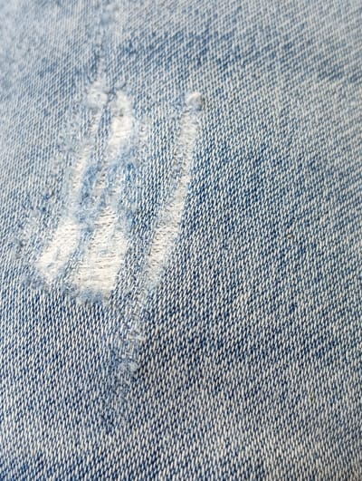 This is a close-up, high-angle shot of worn blue denim fabric. The texture of the woven material is clearly visible, with a mix of indigo and white threads creating a classic denim pattern. A noticeable area of damage or wear is present on the left side of the frame, characterized by frayed threads and lighter, almost white patches where the fabric has been thinned or torn. The lighting appears to be even, suggesting an indoor setting or an overcast day. There are no people, objects, or specific location cues visible in the image, only the abstract texture of the fabric. The time of day, weather, and emotions are not discernible. There is no visible text in the media file. The context of Ruda Śląska, Poland, is provided but does not directly relate to any visual elements within the image itself.