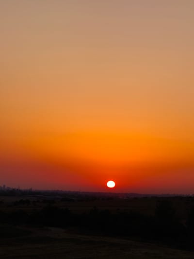 A vertical, low-angle shot captures a dramatic sunset over Sha'alvim, Israel. The sun, a bright white orb, is low on the horizon, casting a fiery orange and red glow across the sky. The landscape below is silhouetted against the vibrant sky, with dark trees and rolling hills creating a stark contrast. In the distance, the faint outline of buildings suggests the presence of a city. The overall impression is one of serene natural beauty, tinged with the subtle presence of human civilization.