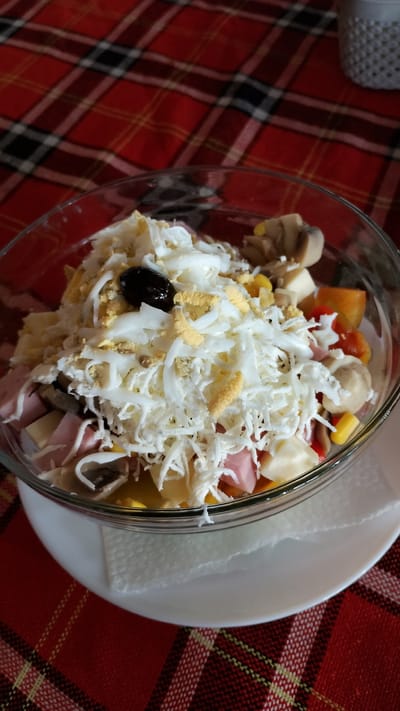 In a glass bowl on a white saucer, a salad is presented. The salad consists of chopped ingredients including what appears to be shredded cheese, hard-boiled egg whites, and egg yolks. Visible among the shredded toppings are diced ham or some type of meat, corn kernels, sliced mushrooms, and a single black olive. The bowl sits on a white napkin which is on a white saucer. The entire setup is placed on a red and black plaid tablecloth. The scene is set indoors, suggested by the artificial lighting and the tablecloth. The location is described as Pevtsite, Bulgaria.