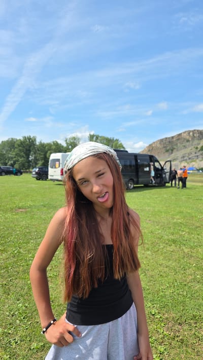A young girl with long, reddish-brown hair and a bandana on her head sticks her tongue out at the camera, with a playful expression. She is wearing a black tank top and gray sweatpants. She has a beaded bracelet on her right wrist. The girl is outdoors in a grassy field with trees and a mountain in the background. Several vans are parked in the field, suggesting a gathering or camping trip. The sky is bright blue with some wispy clouds, indicating it is daytime and likely sunny. The scene appears to be casual and relaxed, possibly a family outing or a recreational activity.