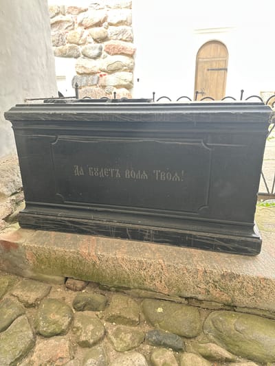 The image shows a dark granite sarcophagus situated on a stone foundation, with cobblestones in the foreground. The sarcophagus has an inscription in Cyrillic letters on its front face, reading "Да будет воля Твоя!" which translates to "Thy will be done!". In the background, a stone wall with large, rough-hewn stones is visible, along with a white building and a wooden arched door. The sarcophagus is located at the Solovetskiy Monastery in Russia. The lighting suggests it is daytime, possibly overcast, as there are no strong shadows. No people are present in the frame.