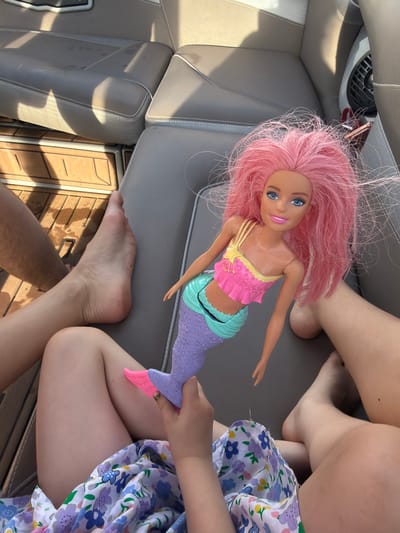 The image depicts the interior of a vehicle, possibly a boat, given the wood paneling. A pink-haired mermaid doll is prominently featured, positioned on the gray upholstered seat. Adjacent to the doll, a child is holding the doll's tail. The child is wearing a floral-patterned dress. An adult's bare foot is visible near the child. The location, Терехово, Russia, is not discernable from the image.