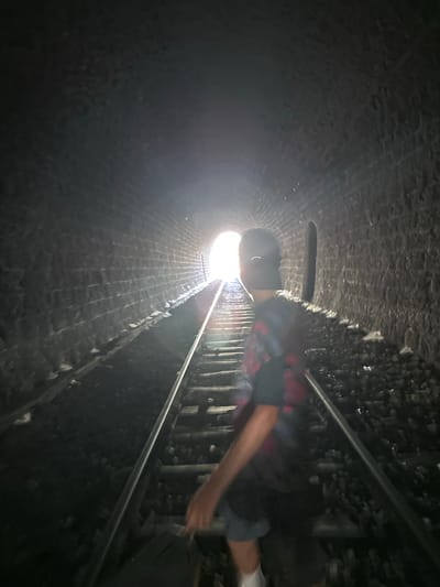 The image shows a person standing on railway tracks inside a stone tunnel. The person is facing away from the camera, towards a bright light at the end of the tunnel. The tunnel walls are made of rough-hewn stones, and the railway tracks are covered in ballast. The scene appears to be located in the Mont Bouquet tunnel in Générargues, France, which is known for its abandoned railway line and has become a popular spot for urban exploration. The person is wearing a t-shirt and shorts, and has a baseball cap on. The lighting is dim, with the bright light at the end of the tunnel creating a high contrast.