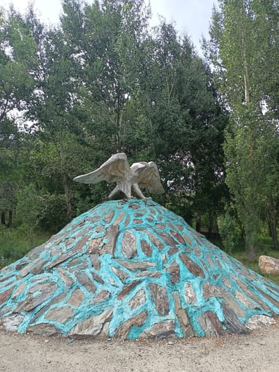 An outdoor, eye-level, medium shot captures a stone sculpture of an eagle with outstretched wings, perched atop a conical mound. The mound is covered with irregular, flat stones, some of which are painted with bright turquoise grout, creating a mosaic-like effect. The eagle sculpture, made of a lighter colored material, is detailed, with visible feathers and talons. Behind the sculpture, a dense backdrop of lush green trees dominates the scene, with a glimpse of a pale, overcast sky peeking through the canopy. The ground in the foreground appears to be a mixture of dirt and gravel. The location is identified as Izgutty Ajtykov, Kazakhstan.