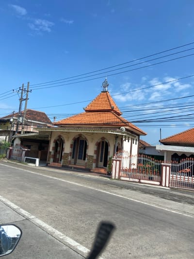 The image shows a small mosque with an orange roof, located on a paved road in the village of Tulangan, Sidoarjo, Indonesia. The mosque is built in a traditional style with arched windows and a decorative gate. The mosque is surrounded by houses and lush greenery. The sky is clear and blue. It is a sunny day, and there are no people present in the image. The mosque is located on the left side of the road, with a white painted line marking the edge of the road. There are several power lines running across the sky. The mosque has an ornately decorated roof. There is a second, smaller house to the right of the mosque, with a similar style to the mosque, also featuring a red tile roof.  The road is paved with grey asphalt and is empty except for a motorcycle in the foreground. There are tall palm trees and green vegetation in the background. The mosque is visible from the road and is a prominent landmark in the area.